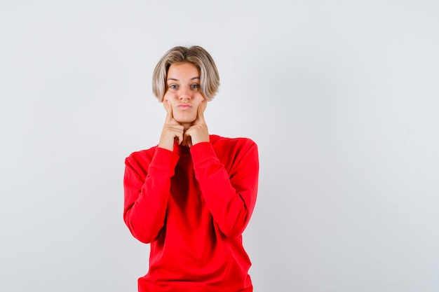 Young teen boy holding fingers on cheeks in red sweater and looking gloomy. front view.