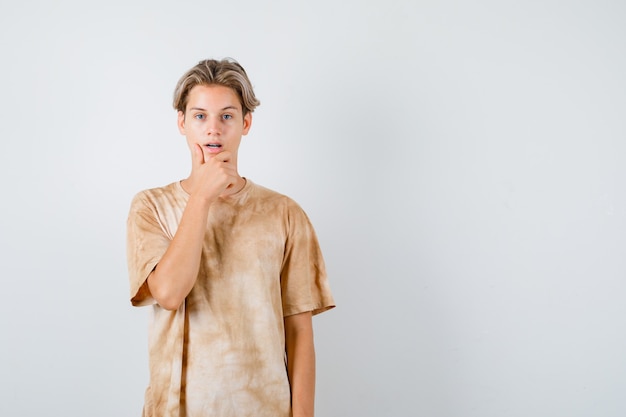 Young teen boy keeping hand on chin in t-shirt and looking pensive. front view.