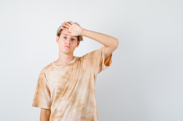 Young teen boy keeping hand on forehead in t-shirt and looking disappointed. front view.