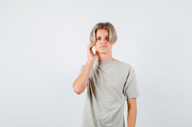 Young teen boy keeping hand near ear in t-shirt and looking confused , front view.