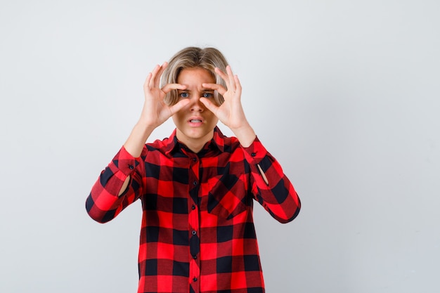 Young teen boy looking through fingers in checked shirt and looking focused , front view.