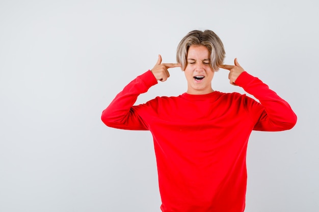 Photo young teen boy plugging ears with fingers in red sweater and looking irritated. front view.