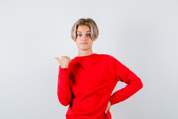 Young teen boy pointing left with thumb in red sweater and looking indecisive , front view.