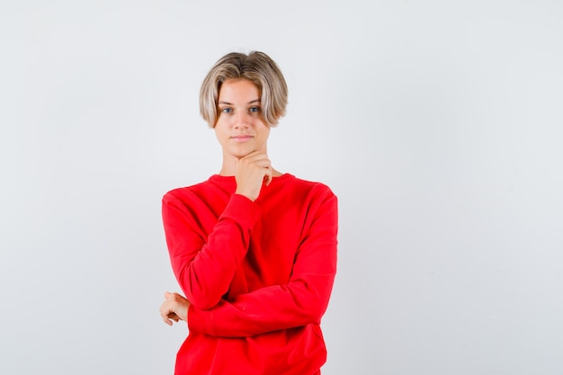 Young teen boy propping chin on hand in red sweater and looking cheery , front view.