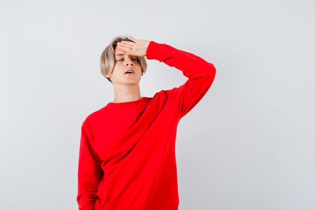 Young teen boy in red sweater with hand on forehead and looking fatigued , front view.