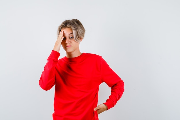 Young teen boy in red sweater with hand on head, looking down and looking thoughtful , front view.