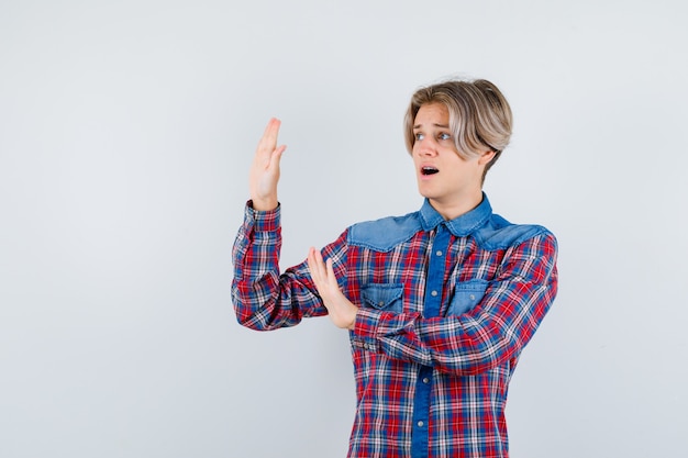 Young teen boy showing stop gesture in checked shirt and looking scared , front view.