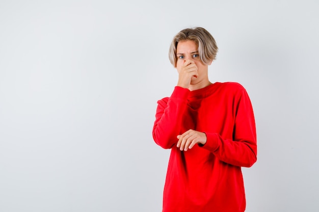 Giovane ragazzo adolescente che odora qualcosa di terribile, pizzicando il naso in un maglione rosso e guardando disgustato, vista frontale.