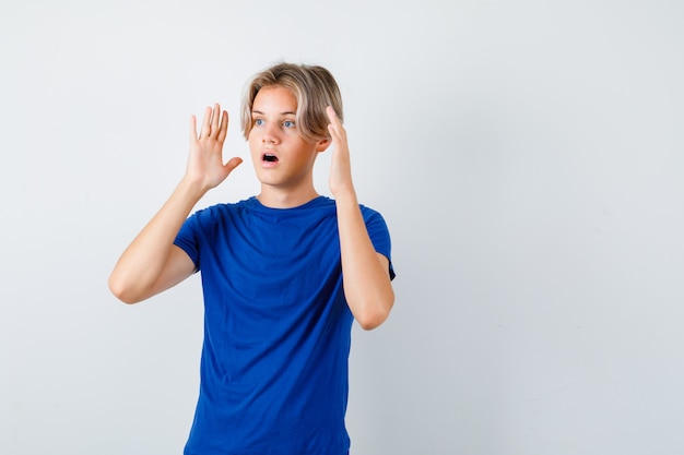 Young teen boy with hands near head while looking away in blue t-shirt and looking shocked. front view.