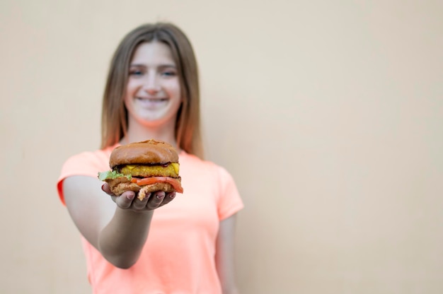 若い十代の少女は、オレンジ色の T シャツを着てオレンジ色の壁に立ち、大きなハンバーガーを保持しています。