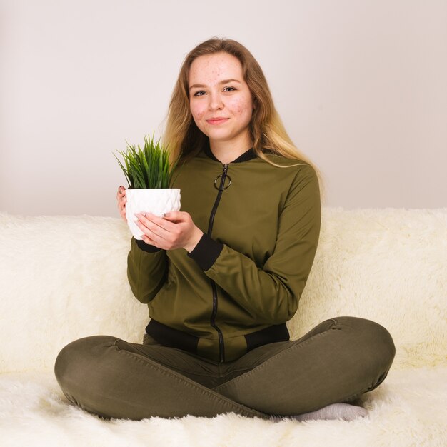 Young teen girl with acne face sitting on sofa and holding green houseplant - image