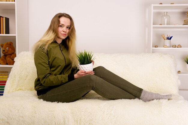 Young teen girl with acne face sitting on sofa and holding green houseplant -