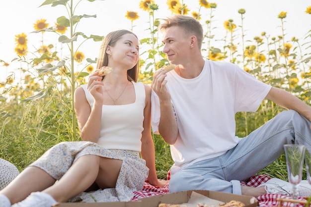 Young teenage couple in white shirts having picnic on sunflower field in sunset. Eating pizza and drinking champagne