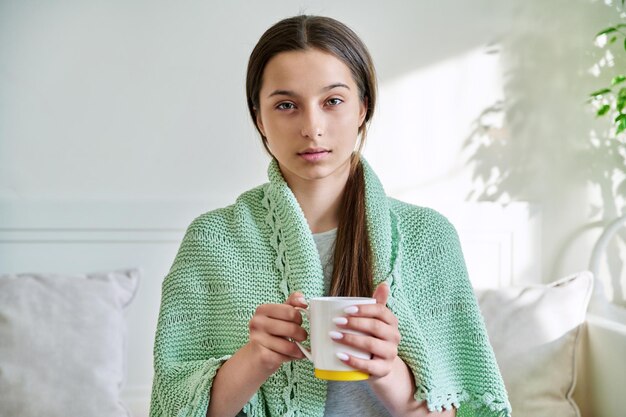 Young teenage female under knitted plaid warming up with hot tea in mug looking at camera