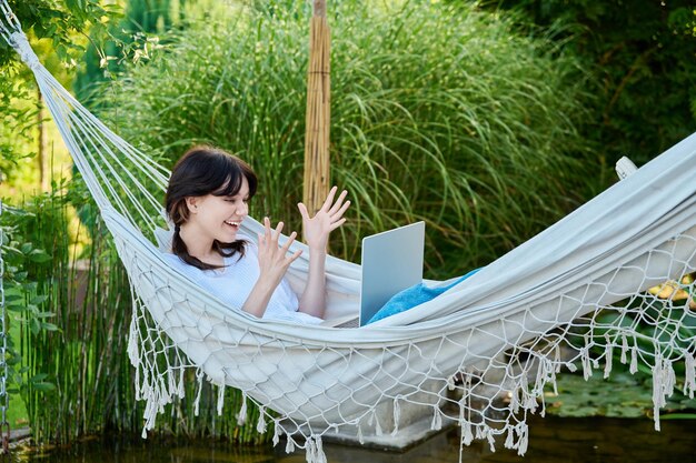 Young teenage female lying outdoor in hammock with laptop talking with friends relatives via video call Happy girl 18 19 years old relaxing talking online using webcam at conference