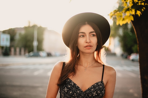 Young teenage girl in a white dress and hat. style of the big city. Sunset sky and rays of the sun. Natural beauty pretty woman in black fedora hipster hat, on warm summer evening sunset in big city