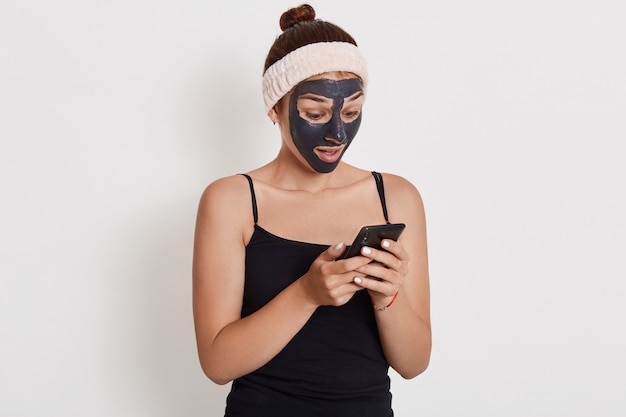 Молодая девушка с черной маской на лице с помощью своего смартфона, глядя на экран устройства с удивленным выражением лица, девушка делает косметические процедуры.