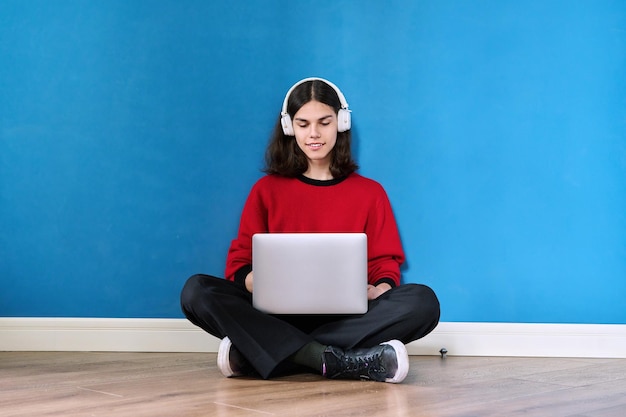 Young teenage guy student in headphones with laptop sitting on the floor on blue background Lifestyle youth technology leisure music education young people concept