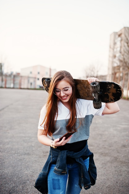 Giovane ragazza urbana adolescente con skateboard, indossare occhiali, berretto e jeans strappati al campo sportivo del cortile sul tramonto facendo selfie sul telefono.