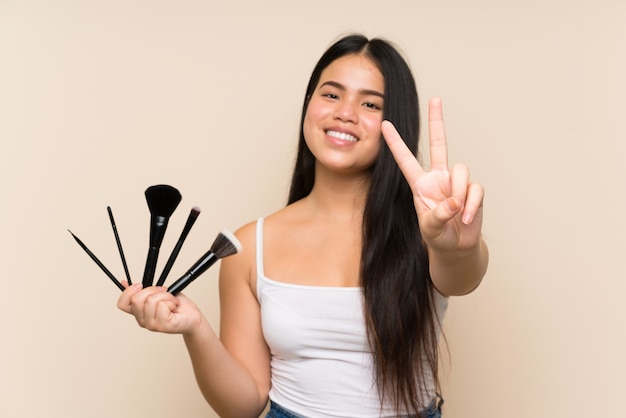 Young teenager Asian girl holding a lot of makeup brush smiling and showing victory sign