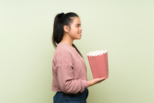 ポップコーンのボウルを保持している孤立した緑の背景の上の若い10代のアジアの女の子