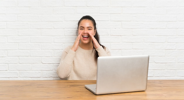 Young teenager Asian girl with a laptop in a table shouting with mouth wide open