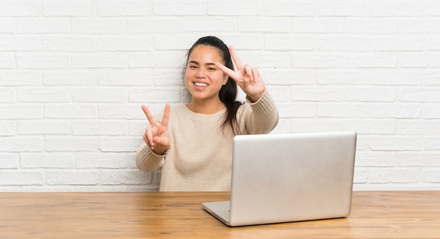 Young teenager Asian girl with a laptop in a table smiling and showing victory sign