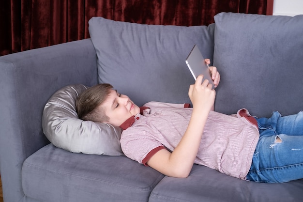 Young teenager boy using tablet lying on grey sofa at home