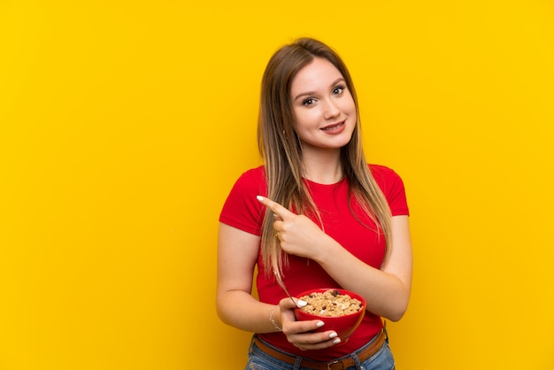 Young teenager girl holding a bowl of cereals pointing to the side to present a product