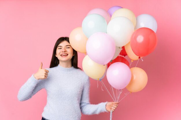 Young  teenager girl holding lots of balloons over pink wall giving a thumbs up gesture