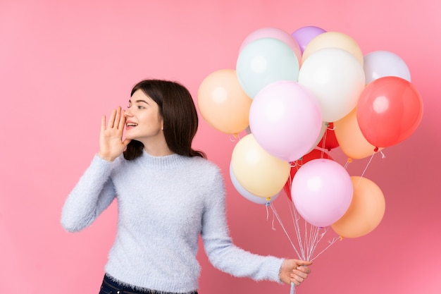 Young  teenager girl holding lots of balloons over pink wall shouting with mouth wide open