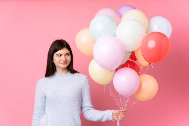 Young  teenager girl holding lots of balloons over pink wall standing and looking to the side