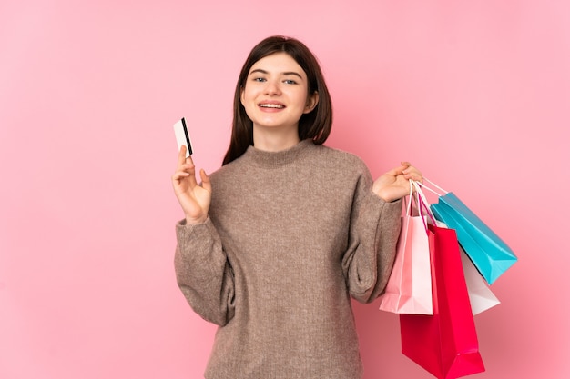 Young teenager girl over  pink wall holding shopping bags and a credit card