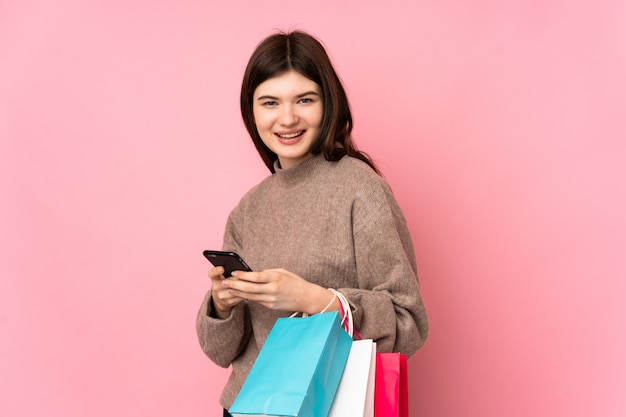 Young  teenager girl over pink wall holding shopping bags and writing a message with her cell phone