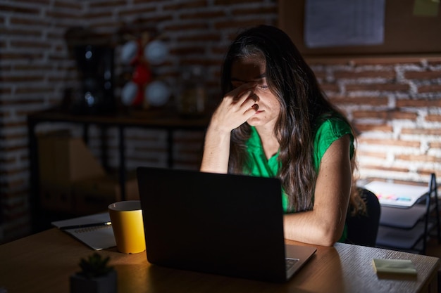 夜にオフィスで働く若い10代の女の子は、鼻と目をこすり、疲労と頭痛を感じて疲れています。ストレスと欲求不満の概念。