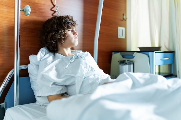 Young teenager sitting in a hospital bed in a ward. Boy preparing for surgery. Sad  depressed child looking aside.  Urgent hospitalization.