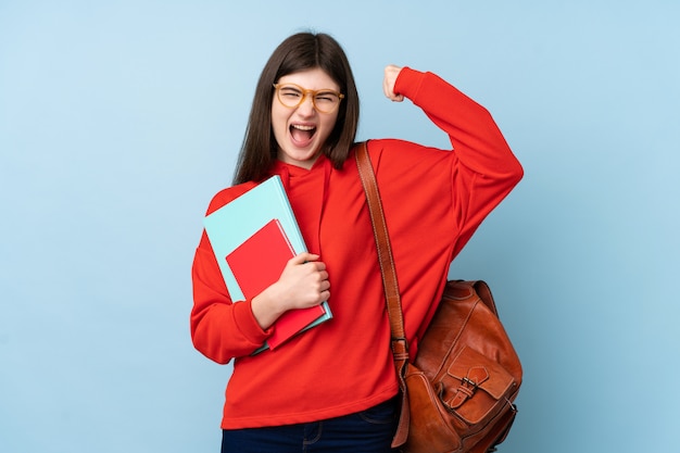 勝利を祝っている青い壁に本を置く若い10代の学生の女の子