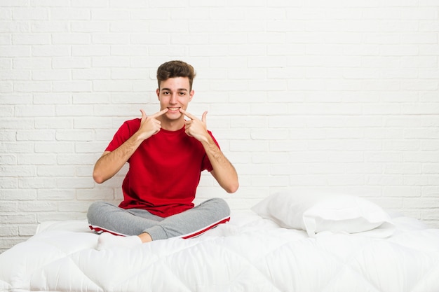 Photo young teenager student man on the bed smiles, pointing fingers at mouth.