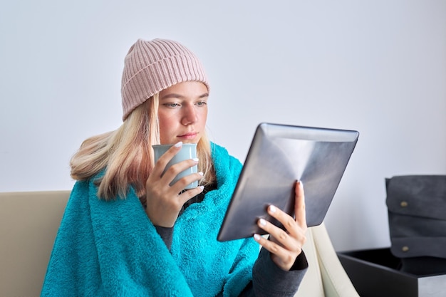 Молодой студент-подросток с цифровым планшетом пьет чашку чая в вязаной шапке под теплым одеялом, сидя дома на диване, изучая досуг в зимний сезон