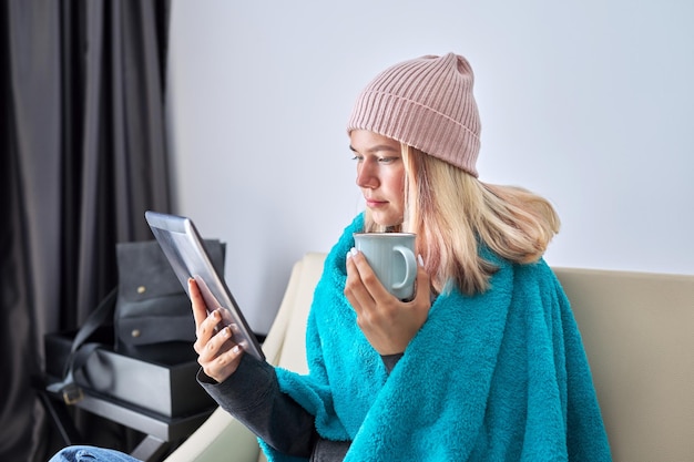 Young teenager student with digital tablet a cup of tea in a knitted hat under a warm blanket sitting at home on the sofa studying leisure lifestyle in winter season