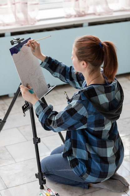 The Young teenager woman artist is sitting on the floor of marble tiles. Woman in the process of painting with oil paints.