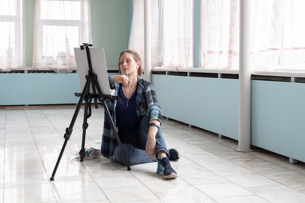 Young teenager woman artist paints with oil paints sitting on the marble floor. White canvas and easel stand on the floor of marble tiles in the room with turquoise and light green walls.