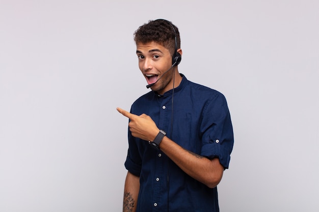 Young telemarketer man looking excited and surprised pointing to the side and upwards to copy space