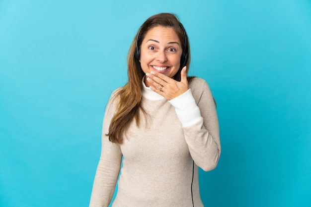 Young telemarketer woman over isolated blue wall happy and smiling covering mouth with hand