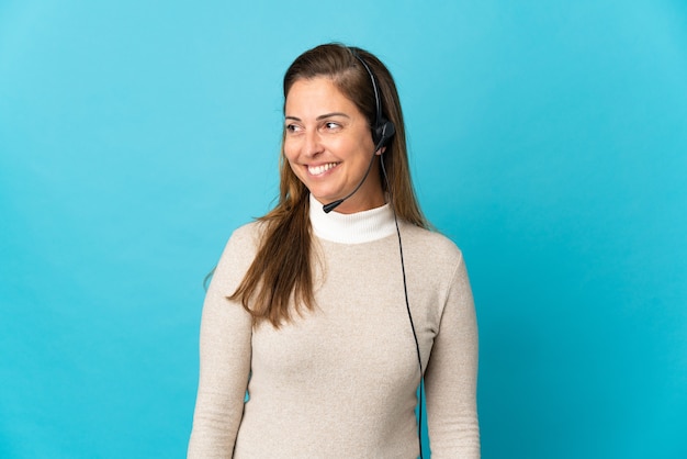 Young telemarketer woman over isolated blue wall looking to the side and smiling