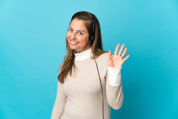 Фото Молодая женщина-телемаркетер над изолированным синим салютом рукой со счастливым выражением лица