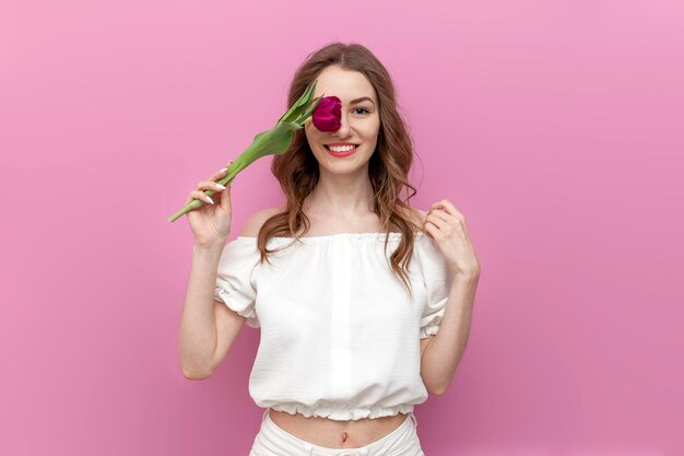 young tender girl closes her eye with pink tulip and smiles on pink isolated background