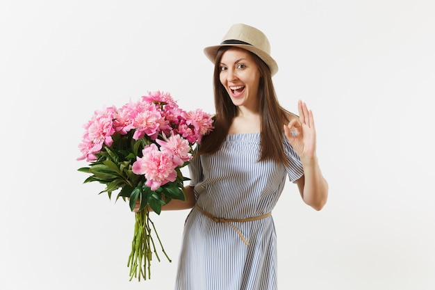Young tender woman in blue dress, hat holding bouquet of beautiful pink peonies flowers, showing OK gesture isolated on white background. St. Valentine's Day, International Women's Day holiday concept