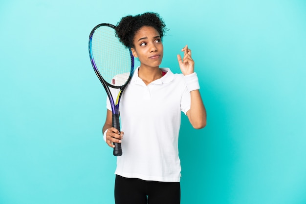 Молодая женщина-теннисистка изолирована на синем фоне со скрещенными пальцами и желает лучшего