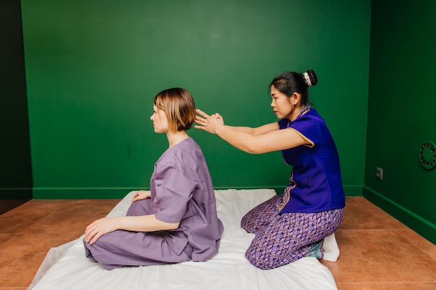 Young Thai masseur worker girl in ethnic asian exotic costume makes and demonstrates different traditional spa procedures  in green relaxation yoga room